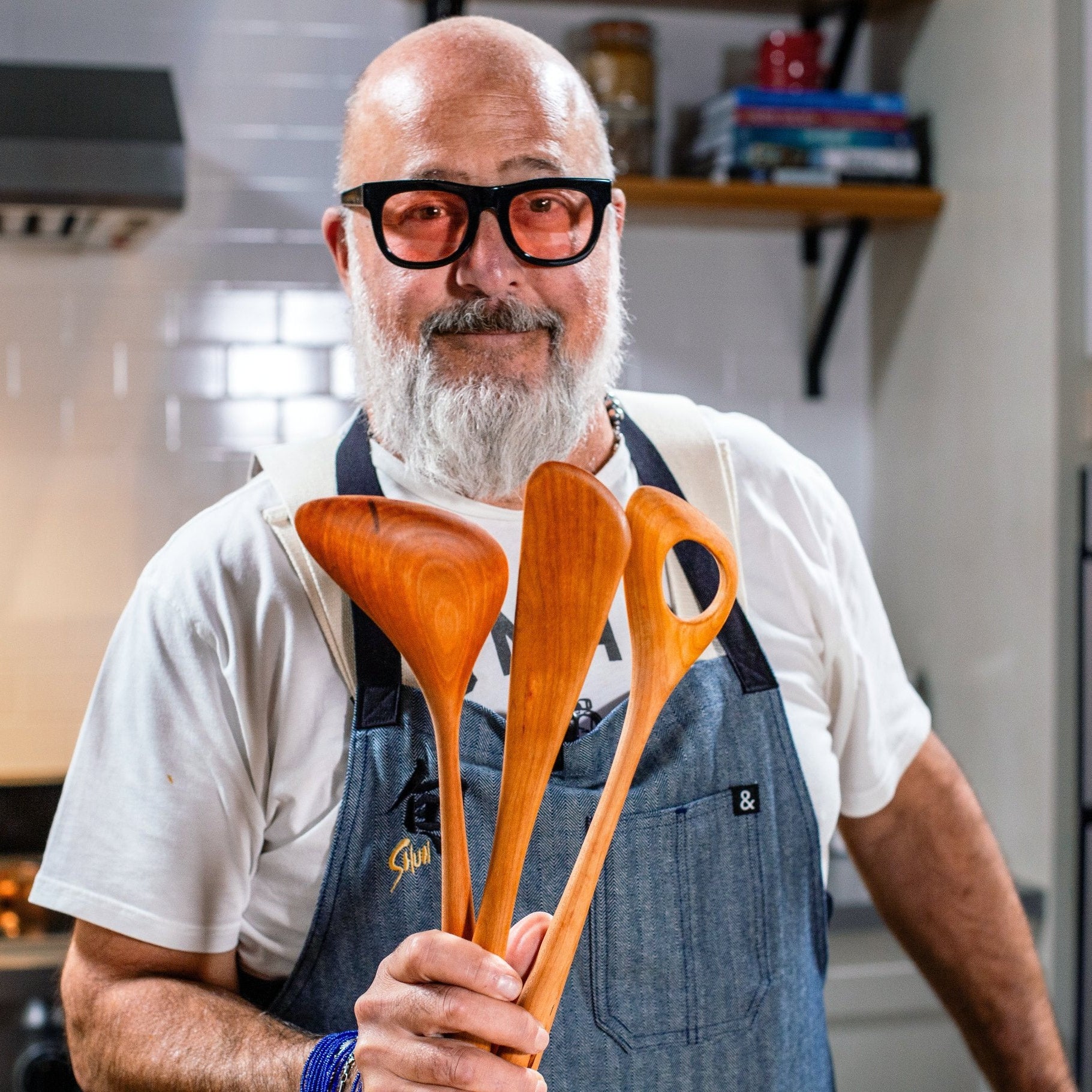 Andrew Zimmern of the Food Network, in a kitchen, smiling and holding wooden spoons and a wooden spatula.