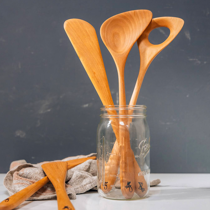 A set of wooden kitchen utensils, including a ladle, spatula, and spoon, are displayed inside and around a glass jar on a countertop. The utensils are crafted from cherry wood and have a polished finish.