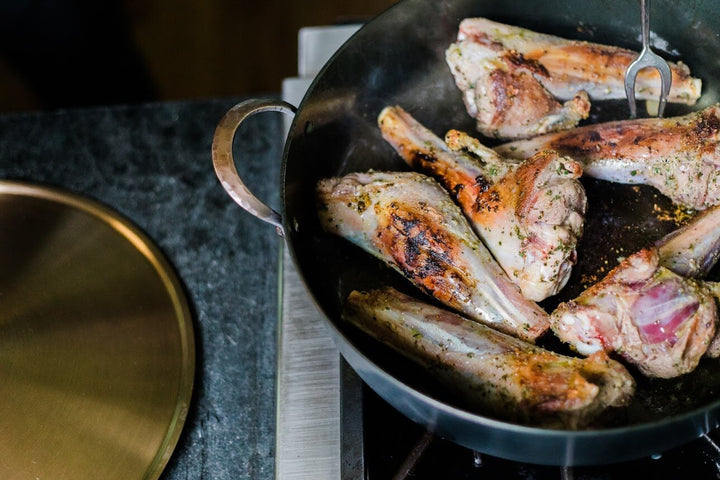 Chicken roasting on a gas stove in a hand forged carbon steel pro rondeau with bronze handles with the stainless steel lid lying next to it