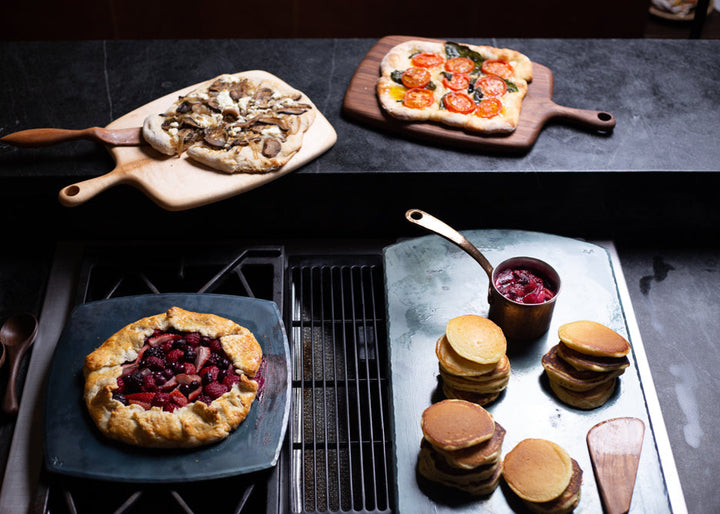 Two handmade carbon steel bakeware sheets with a cooked berry tart and a pile of cooked pancakes and two handmade serving boards with handmade pizzas on top