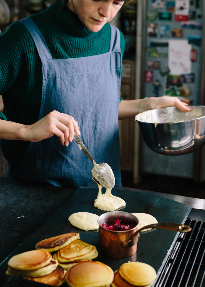 A handcrafted carbon steel bakeware sheet with a woman wearing a bathrobe placing pancake batter on top next to a cooked pile of pancake and a copper cup on jam