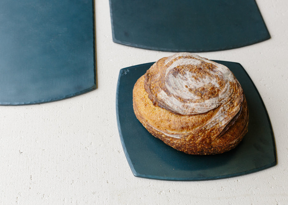 A set of hand forged carbon steel bakeware sheets with one flat on the table and a fresh loaf of bed resting on top