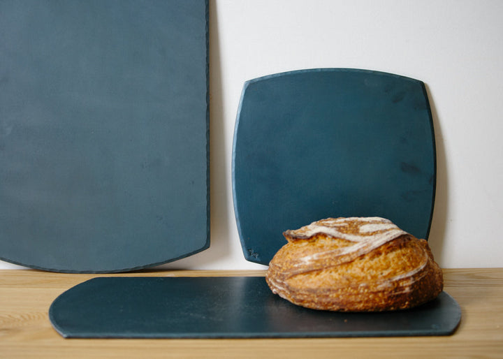 A set of hand forged carbon steel bakeware sheets with one flat on the table and a fresh loaf of bed resting on top