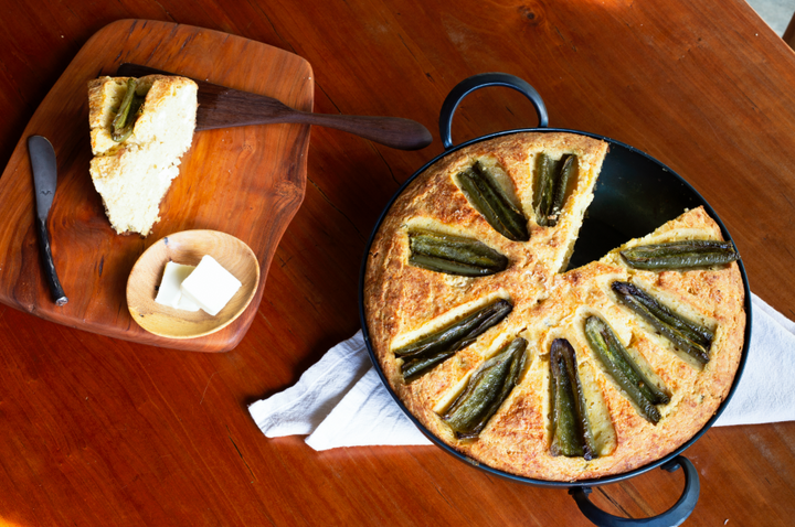 A hand forged, handmade eleven inch saucier holding baked cornbread next to a serving board with a hand forged spreader and hand carved spatula