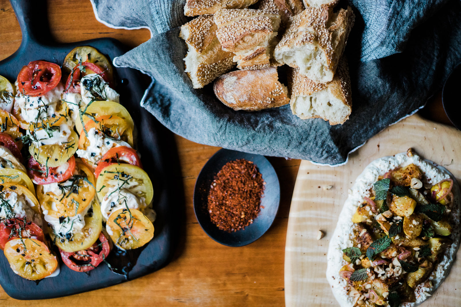 Two handmade wooden serving boards with caprese and handmade pizza atop next to a bowl of bread and a pinch bowl of spice