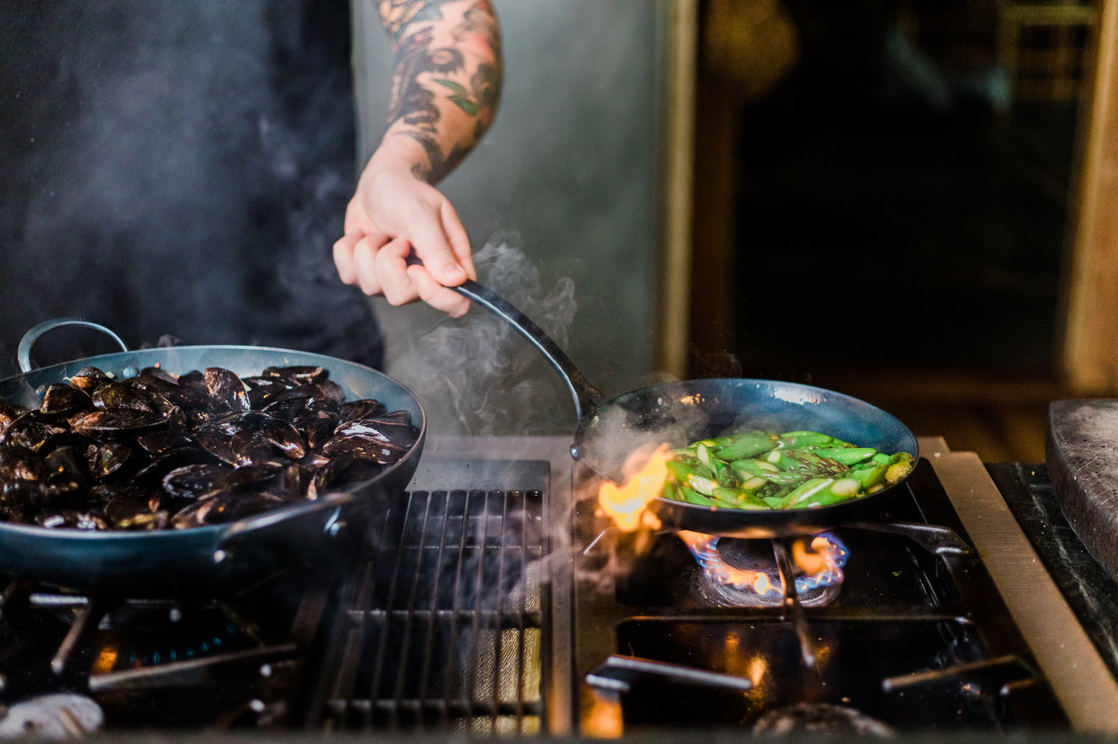 A handmade carbon steel saucier atop a gas stove and a chef cooking green peppers in a hand forged carbon steel skillet