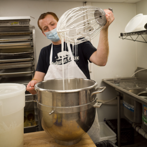 Baker lifting large whisk with cinnamon roll glaze dripping off out of a mixing bowl in a bakery kitchen 