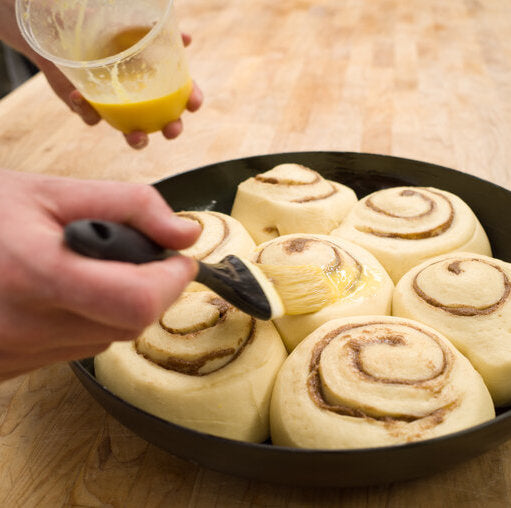 Baker brushing butter onto uncooked cinnamon rolls in a carbon steel saucier pan