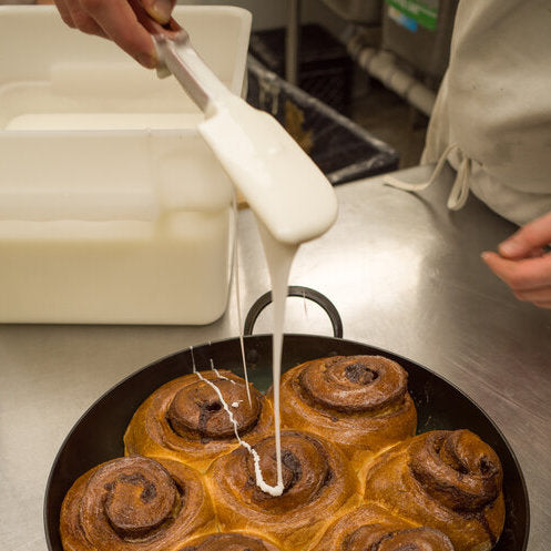 Baker drizzling glaze onto cinnamon rolls in a carbon steel saucier