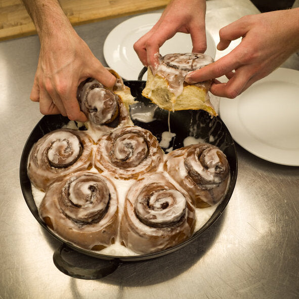 Hands pulling apart a pan of cinnamon rolls in a carbon steel skillet next to two empty plates