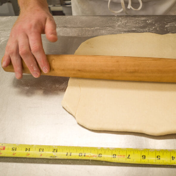Baker rolling out dough next to a measuring tape on a stainless steel surface