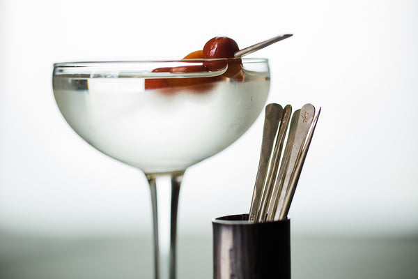 A cocktail glass with a hand forged cocktail pick holding cherries and a cocktail pick container with cocktail picks inside