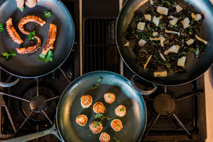 Three hand forged carbon steel pans with a variety of foods cooking on them