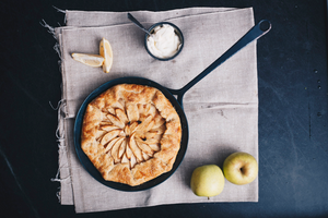 Rustic apple galette in a carbon steel skillet pan lying on a linen napkin with whole apples, lemon slices and a bowl of cream next to the pan on the napkin