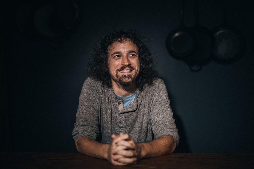 Man with curly hair and beard sitting at a table in a dimly lit room.