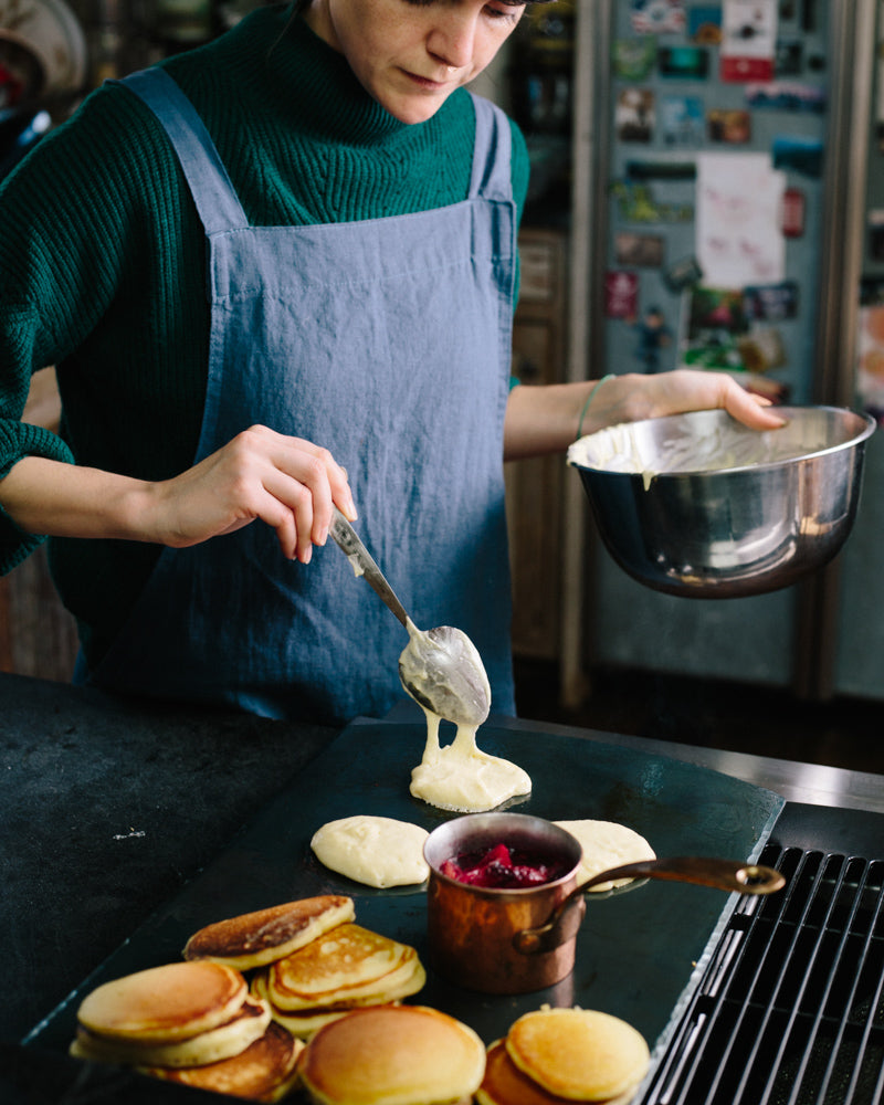 A handcrafted carbon steel bakeware sheet with a woman wearing a bathrobe placing pancake batter on top next to a cooked pile of pancake and a copper cup on jam