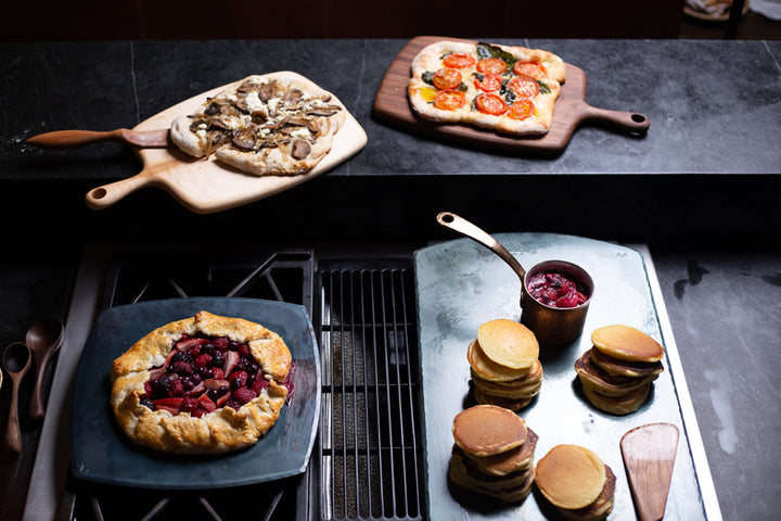 Two handmade carbon steel bakeware sheets with a cooked berry tart and a pile of cooked pancakes and two handmade serving boards with handmade pizzas on top