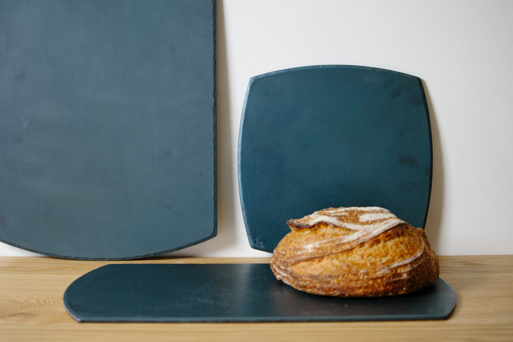 A set of hand forged carbon steel bakeware sheets with one flat on the table and a fresh loaf of bed resting on top