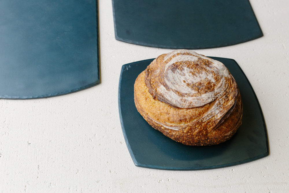 A set of hand forged carbon steel bakeware sheets with one flat on the table and a fresh loaf of bed resting on top