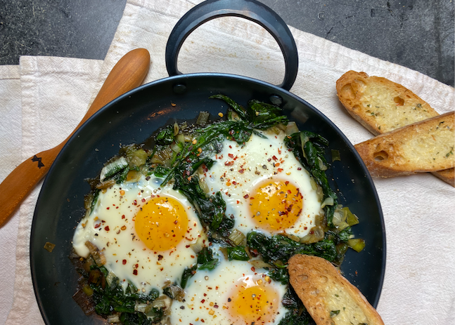 Hand forged nine inch carbon steel pro roaster with fried eggs and collard greens resting on linen napkins with a handmade wooden spreader and two toasted bread slices 