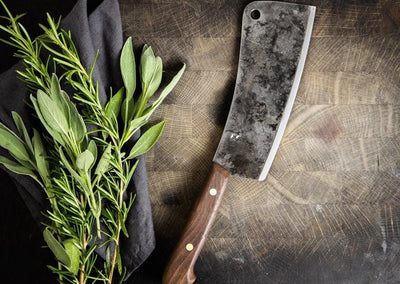 Kitchen cleaver resting on a cutting board next to herbs resting on a napkin