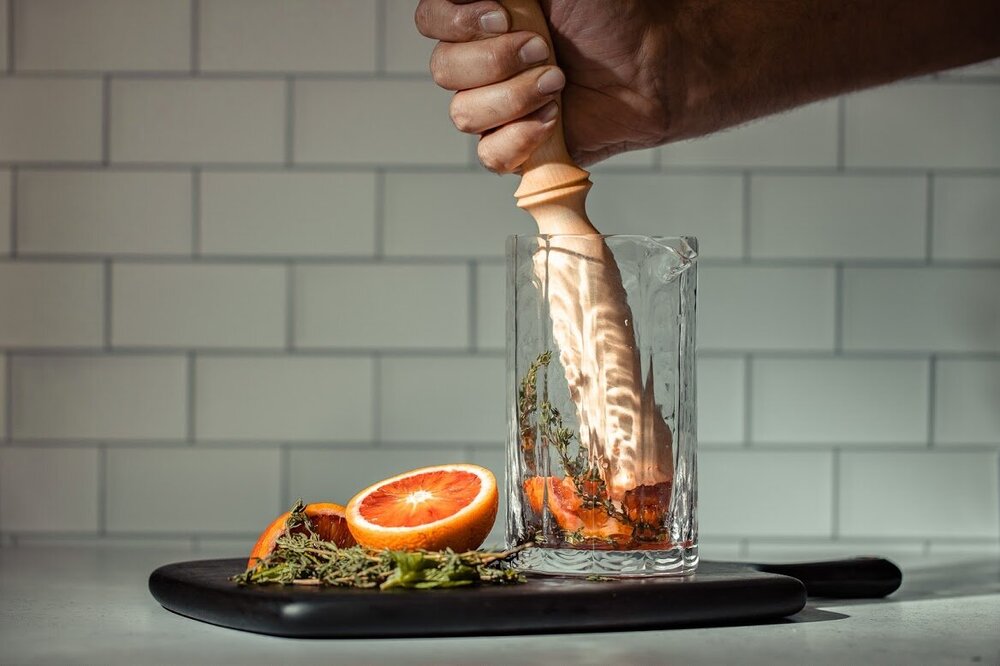 Maple handmade, hand carved cocktail muddler muddling orange and herbs in a cocktail mixer on top of a serving board