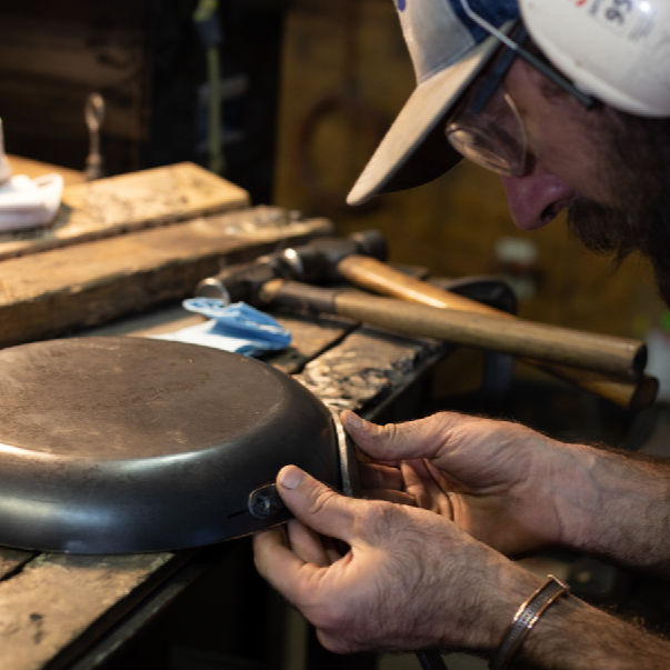 Man forging carbon steel pan in a shop wearing work glasses and protective ear wear