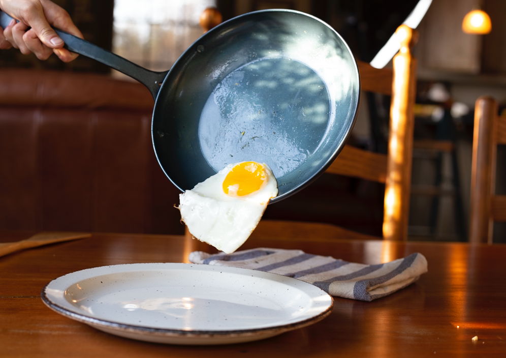 Handcrafted carbon steel pan with a fried egg sliding off it onto a plate below 