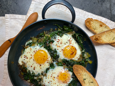 Hand forged nine inch carbon steel pro roaster with fried eggs and collard greens resting on linen napkins with a handmade wooden spreader and two toasted bread slices 