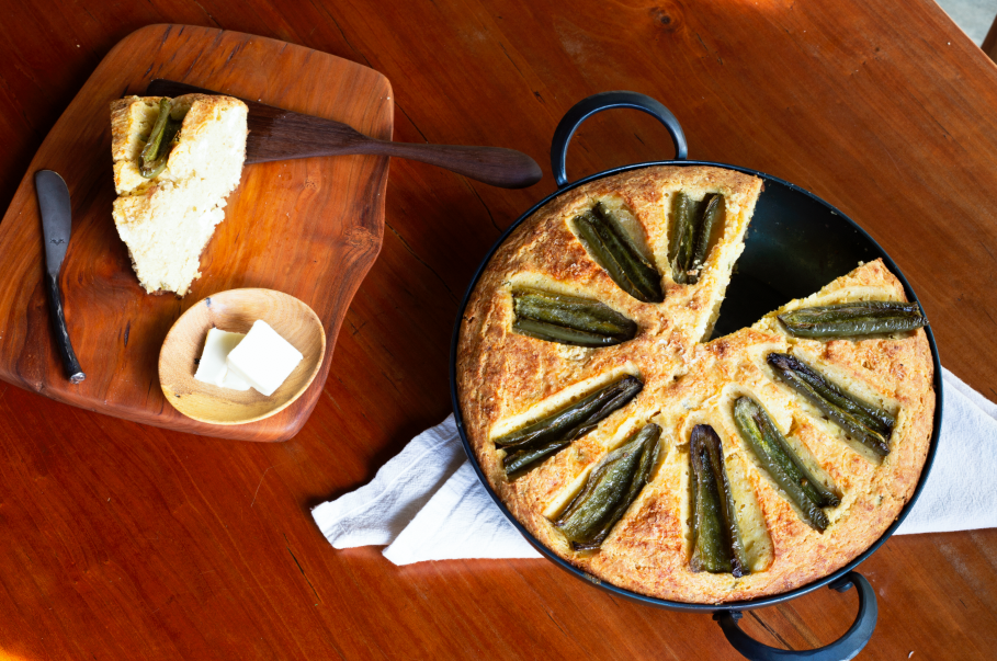 A hand forged, handmade eleven inch saucier holding baked cornbread next to a serving board with a hand forged spreader and hand carved spatula
