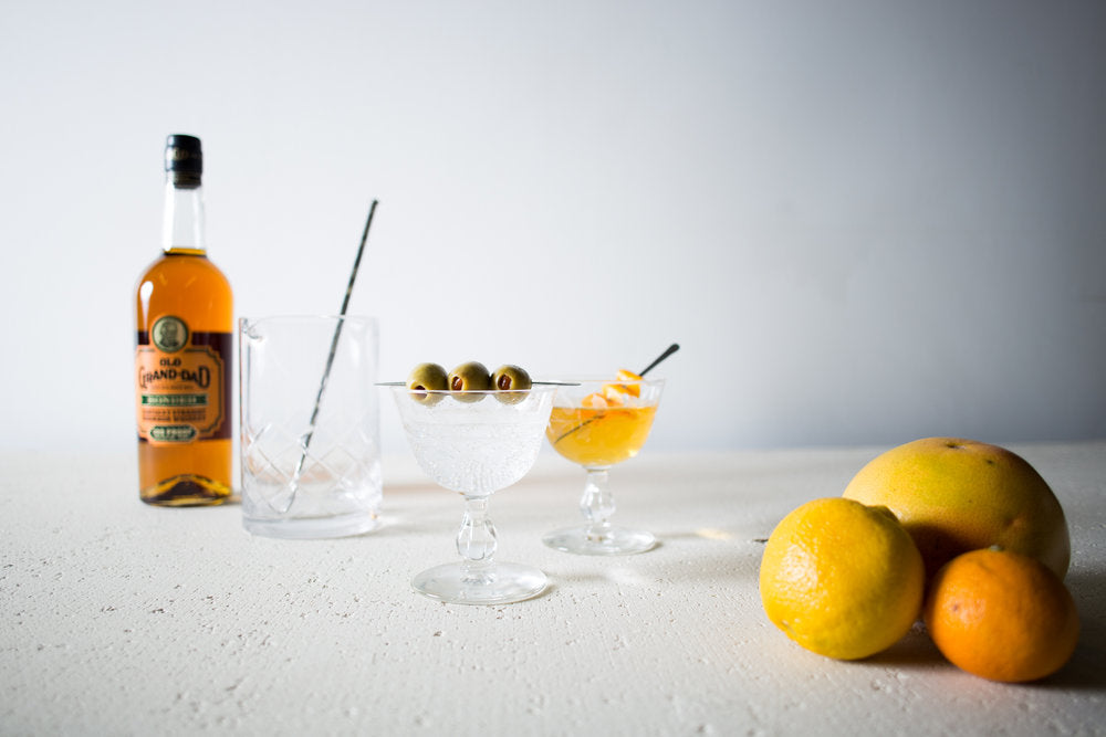 A cocktail spoon in a cocktail mixer glass and two cocktail glasses full of cocktail and handmade artisan cocktail picks holding olives  and orange peels