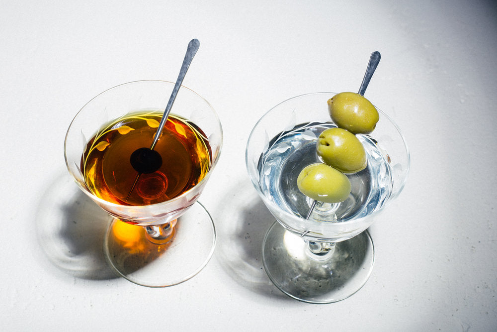 Two cocktail glasses full of cocktails and cocktail picks in the glasses holding a cherry and one pick holding three olives