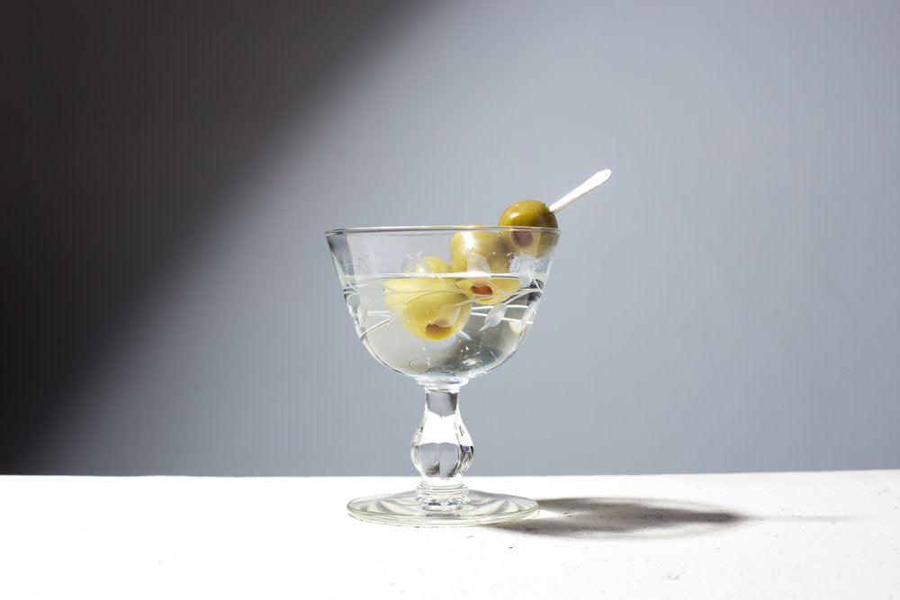 A cocktail glass holding a drink  with a handmade artisan cocktail pick holding three olives 